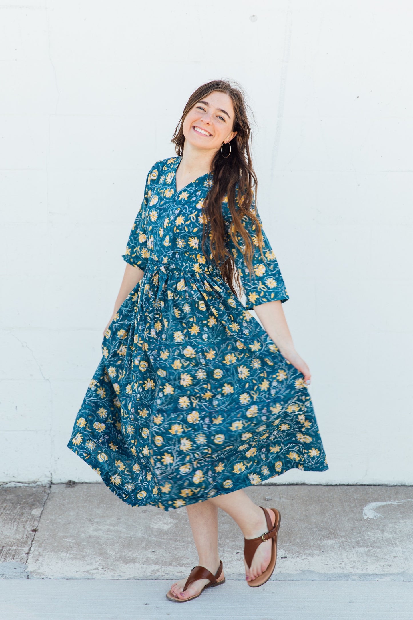 A woman standing and smiling, wearing a blue floral print dress with a v-neckline and 3/4 sleeves.