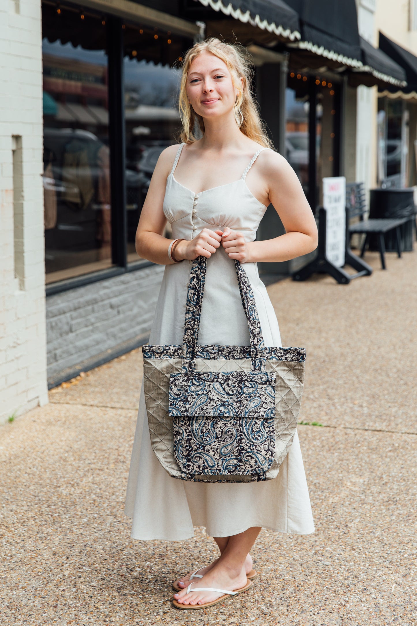 A woman standing on the sidewalk holding a tan tote bage with a blue block-printed quilted design.