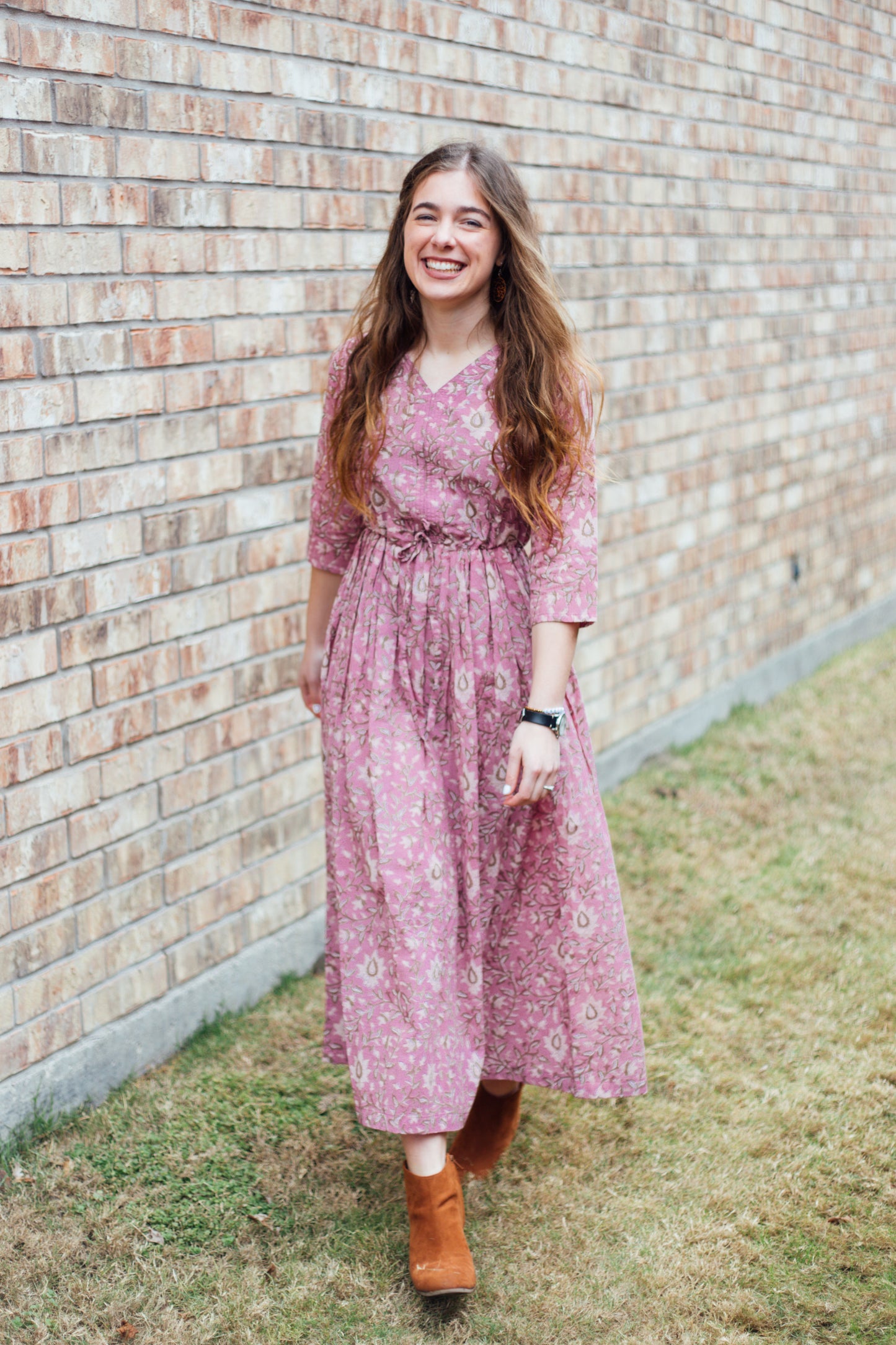 A woman standing and smiling, wearing a pink floral print dress with a v-neckline and 3/4 sleeves.