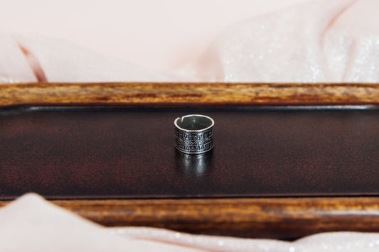 A statement silver ring with intricate patterns placed on a wooden surface with a pink fabric underneath.