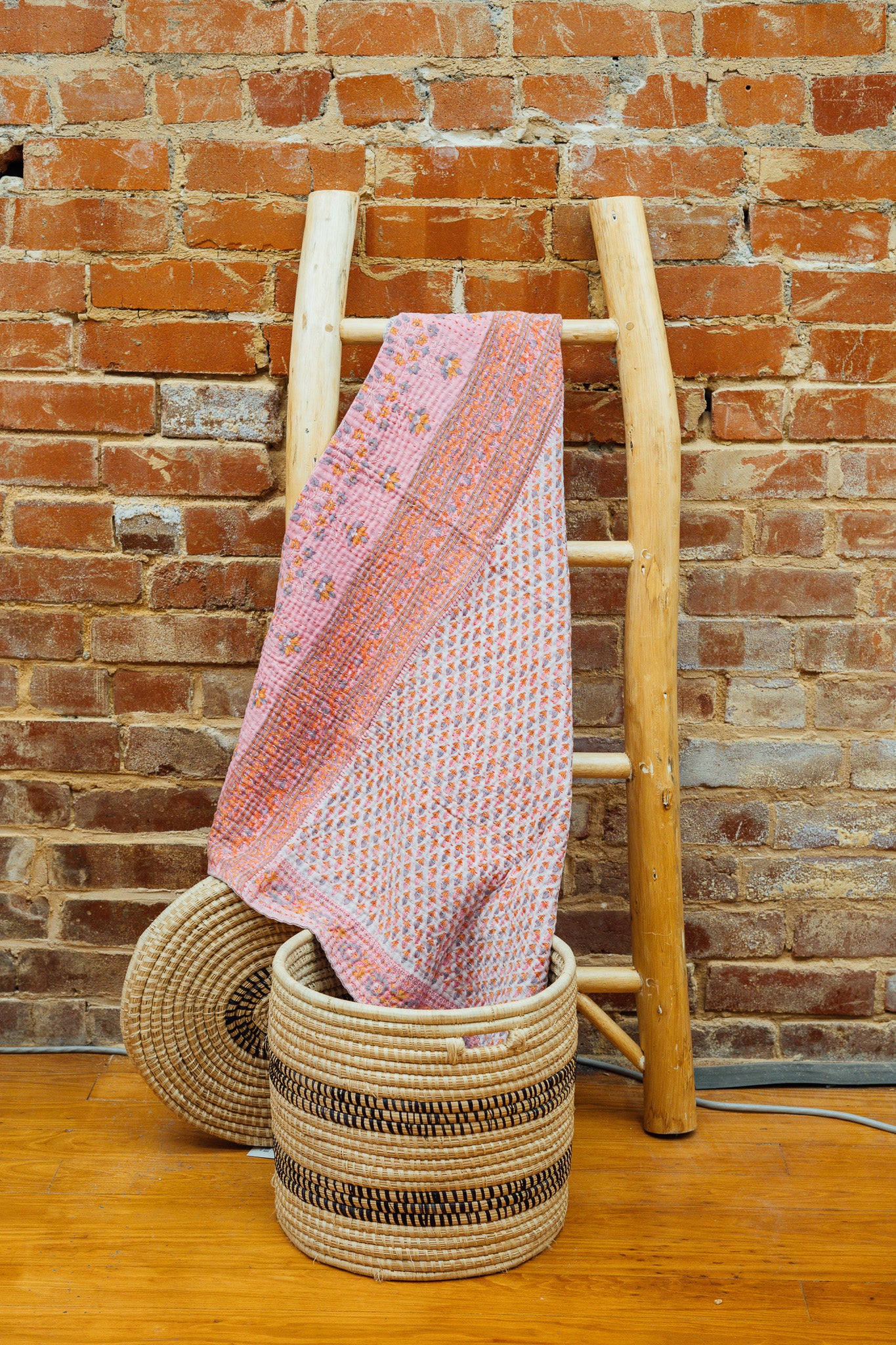 A pink Kantha baby blanket hanging on a wooden rack against a brick wall, with a wicker basket in the foreground.