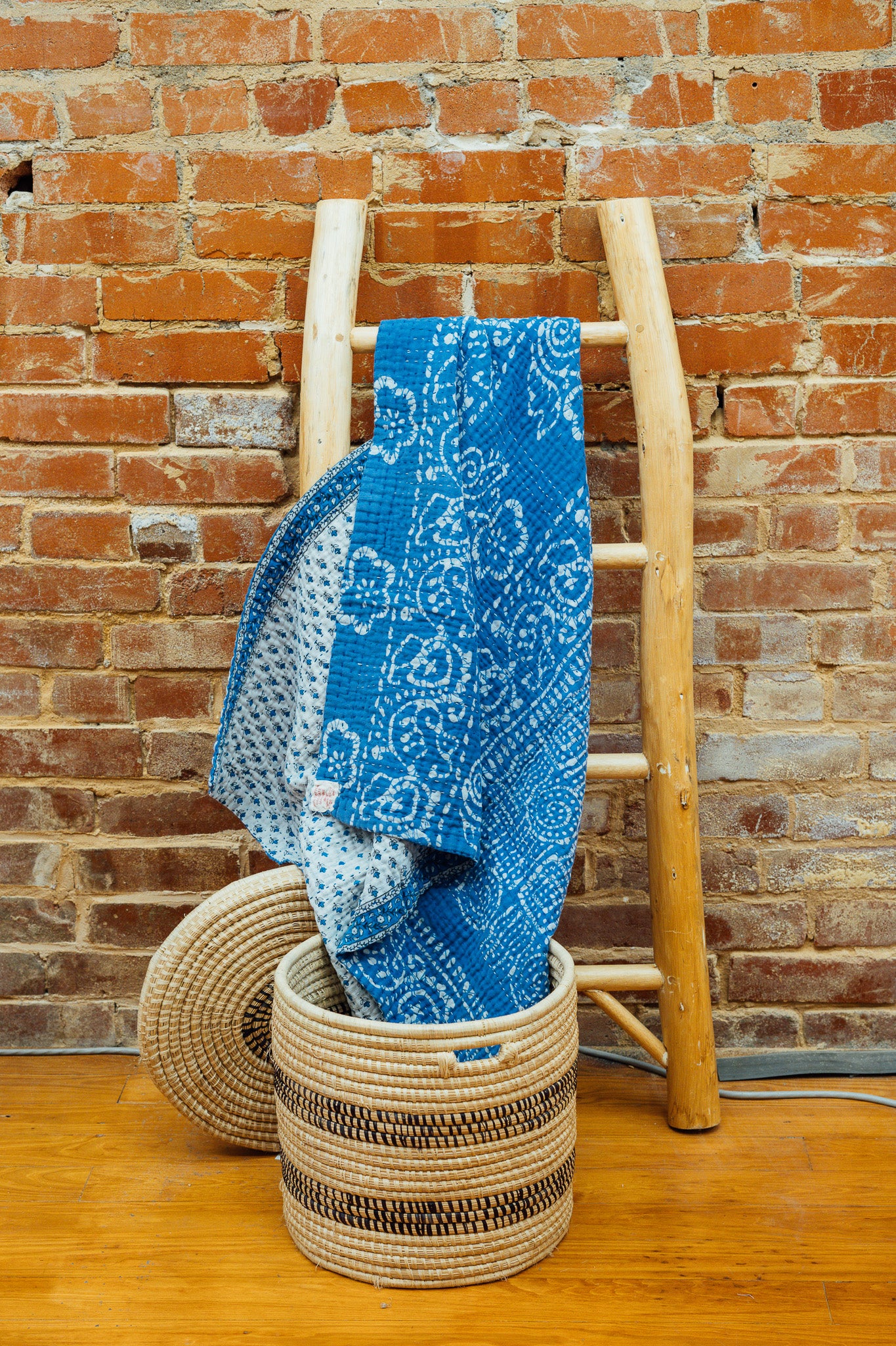 A blue and white Kantha baby blanket hanging on a wooden rack against a brick wall, with a wicker basket in the foreground.