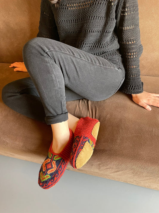 A person sitting on a couch wearing gray pants and red hand-knitted slippers with geometric patterns.