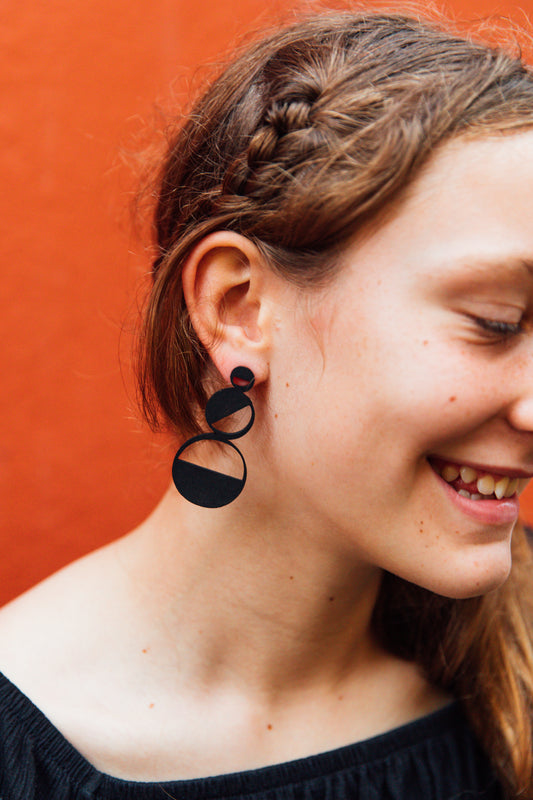 A person wearing a black top and showcasing black wooden earrings against an orange wall.