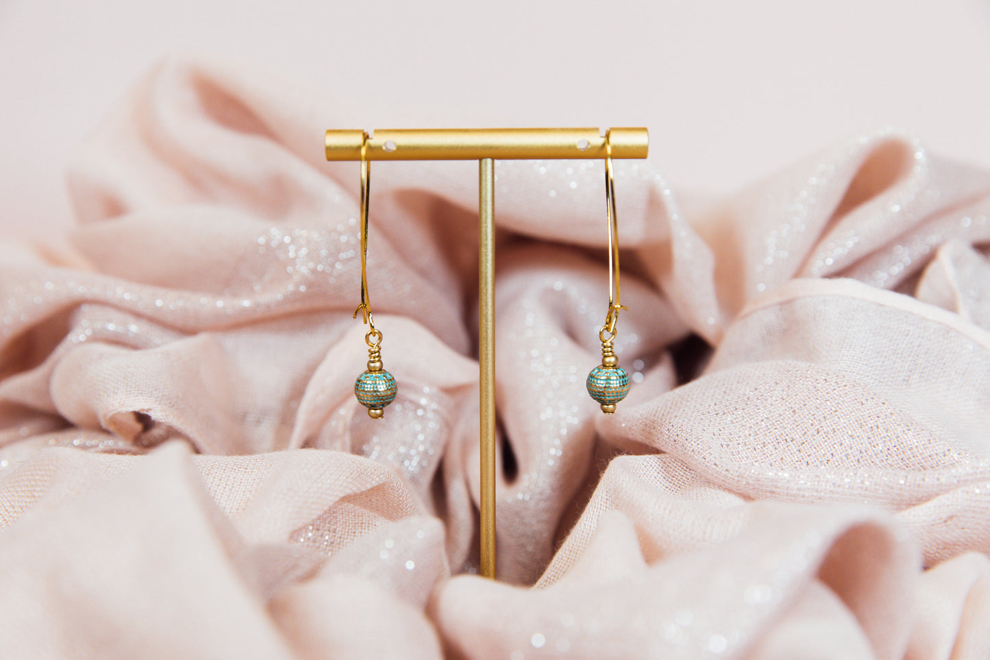 A pair of earrings with gold finished hoops and sea green metal beads, displayed on a pink fabric.