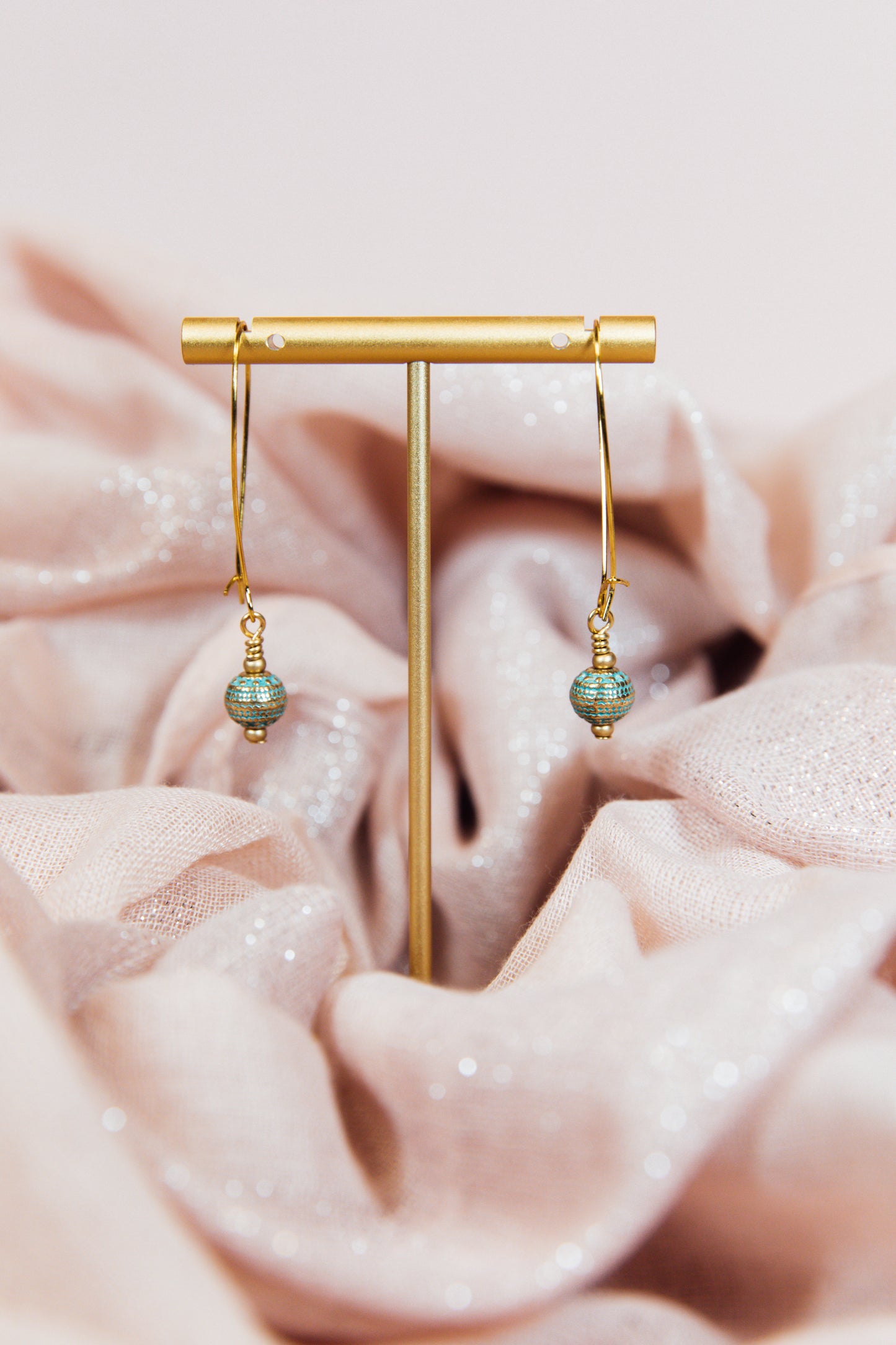 A pair of earrings with gold finished hoops and sea green metal beads, displayed on a pink fabric.