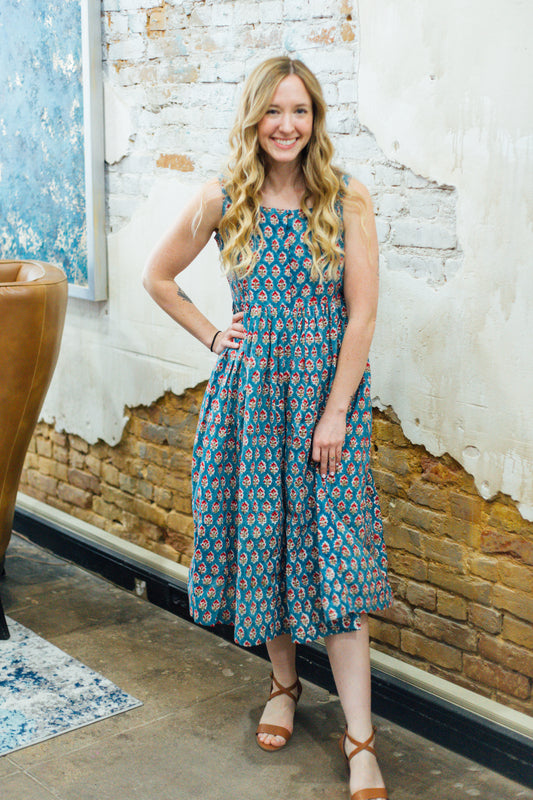A woman standing indoors wearing a blue and red floral dress with three-quarter sleeves and a square-neckline.