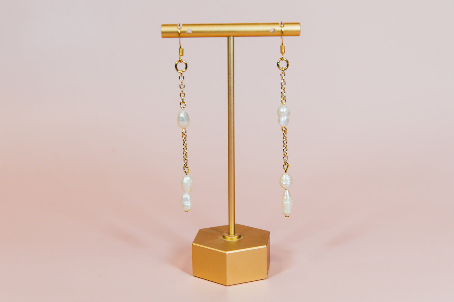 A pair of earrings with fresh water pearls attached to gold-finished chains, displayed on a gold-colored stand against a pink background.