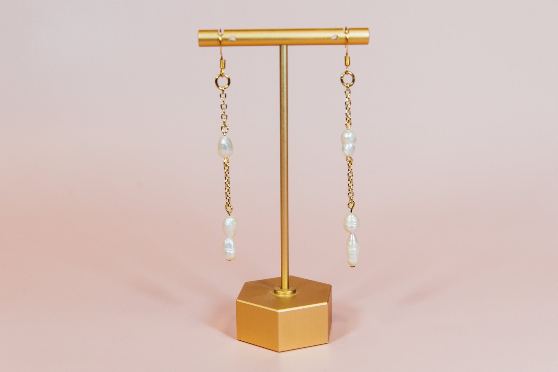 A pair of earrings with fresh water pearls attached to gold-finished chains, displayed on a gold-colored stand against a pink background.