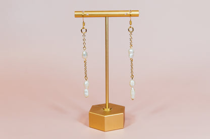 A pair of earrings with fresh water pearls attached to gold-finished chains, displayed on a gold-colored stand against a pink background.