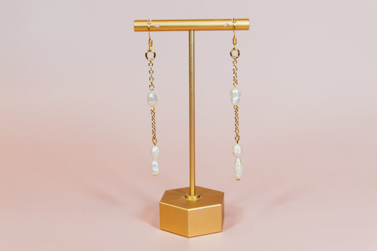 A pair of earrings with fresh water pearls attached to gold-finished chains, displayed on a gold-colored stand against a pink background.
