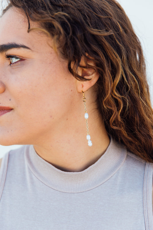 A pair of earrings with fresh water pearls attached to gold-finished chains, displayed on a gold-colored stand against a pink background.