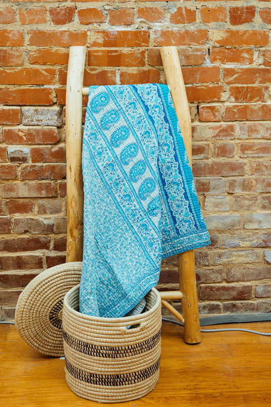 A blue and white Kantha baby blanket hanging on a wooden rack against a brick wall, with a wicker basket in the foreground.