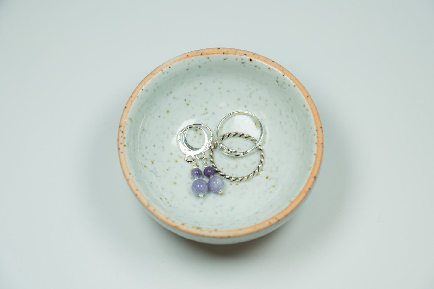 Two silver rings with purple stones in a ceramic dish on a light gray background