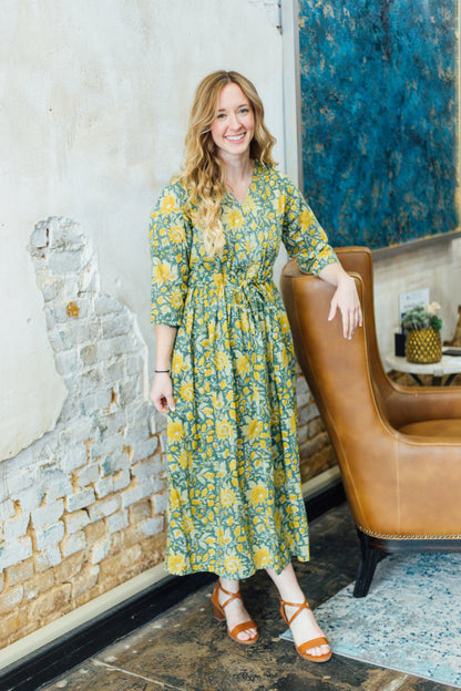 A woman standing and smiling, wearing a green floral print dress with a v-neckline and 3/4 sleeves.