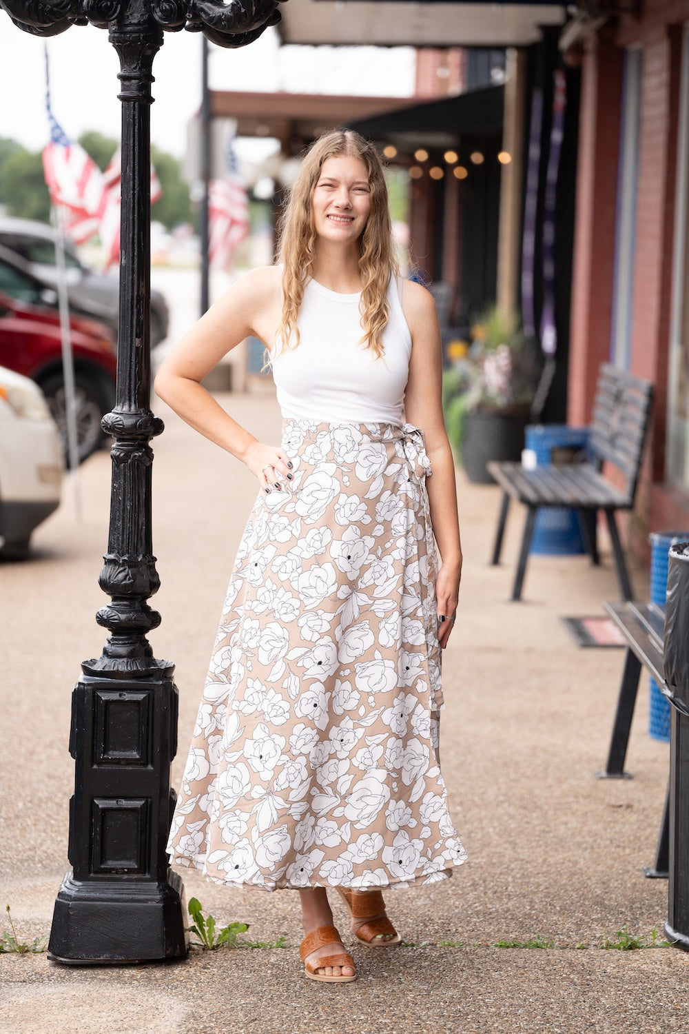 Woman in a white top and floral skirt standing next to a black street lamp on a sidewalk.