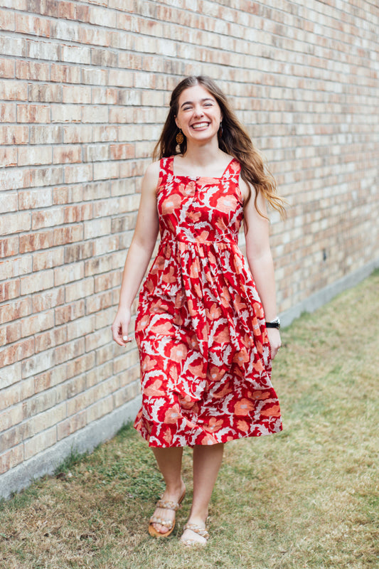 A woman standing outdoors wearing a red and orange floral dress with three-quarter sleeves and a square neckline.