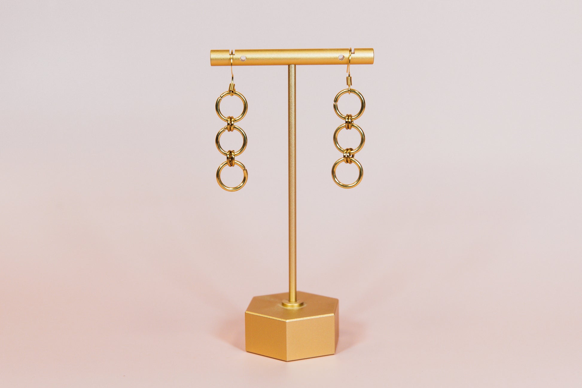 A pair of gold finished triple hoop earrings displayed on a gold-colored stand against a pink background.