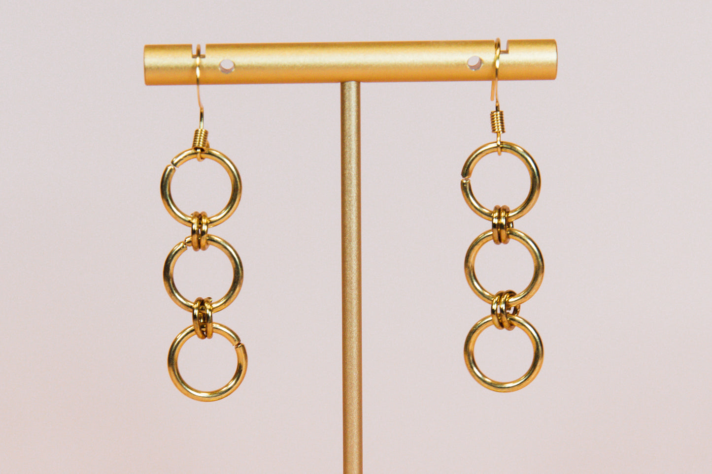 A pair of gold finished triple hoop earrings displayed on a gold-colored stand against a pink background.