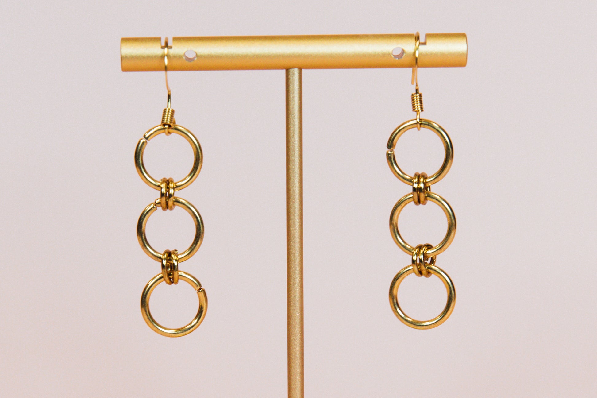 A pair of gold finished triple hoop earrings displayed on a gold-colored stand against a pink background.