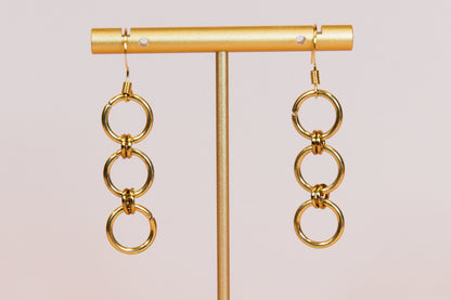 A pair of gold finished triple hoop earrings displayed on a gold-colored stand against a pink background.