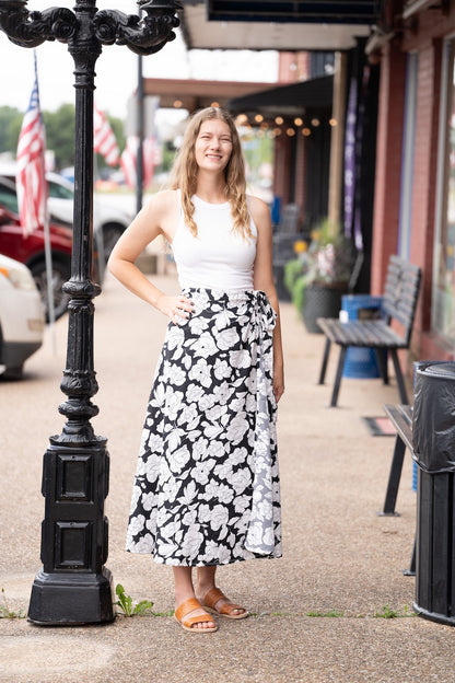 Woman in a white top and black floral skirt standing next to a street lamp on a sidewalk.