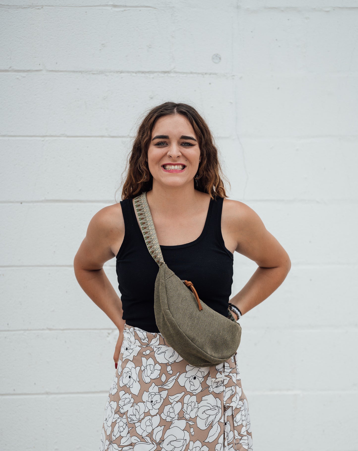 Woman wearing a black tank top and floral skirt with a green Thai Dumpling Sling Bag over her shoulder against a white wall.