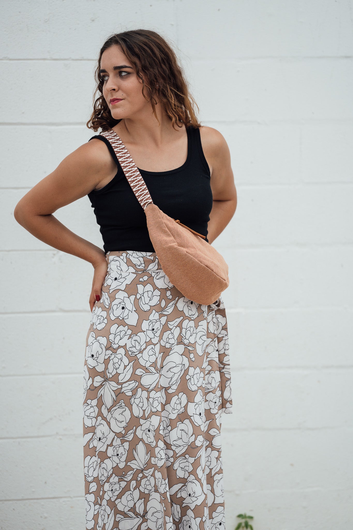 Woman wearing a black top and floral skirt with a Sienna Thai Dumpling Sling bag against a white wall